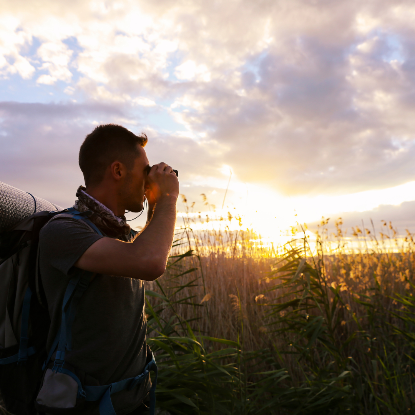 Man with a backpack using Pursual binoculars in a field at sunset